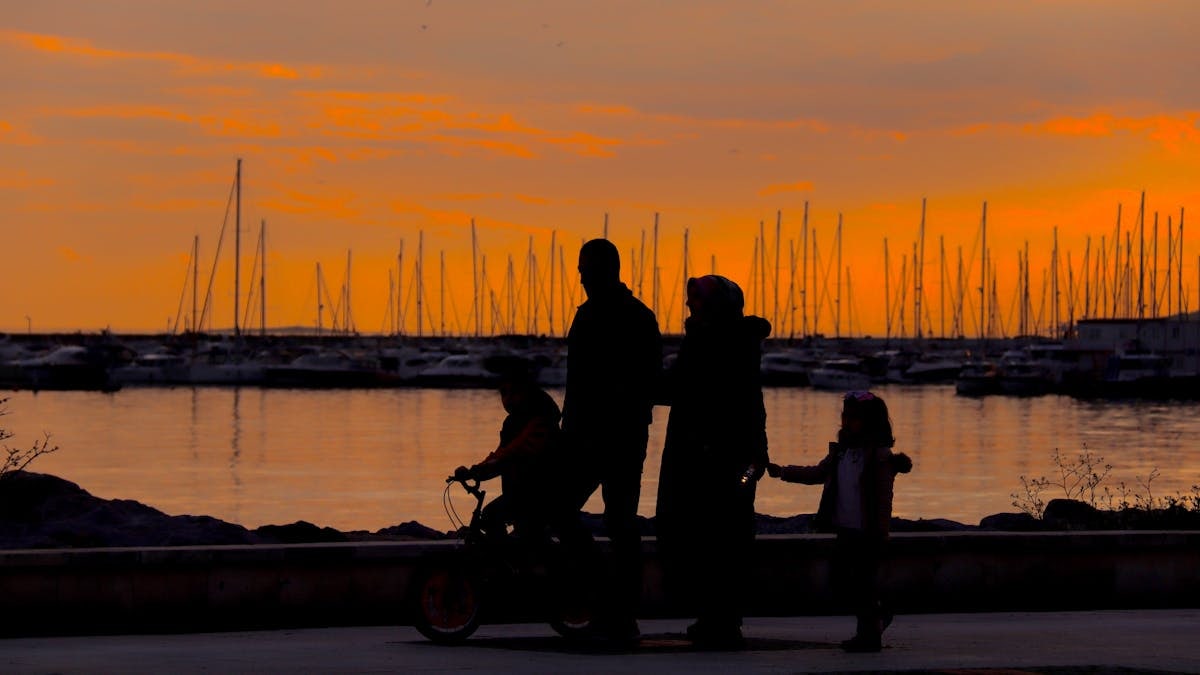 A silhouette of a family walking by the harbor at sunset with boats in the background.