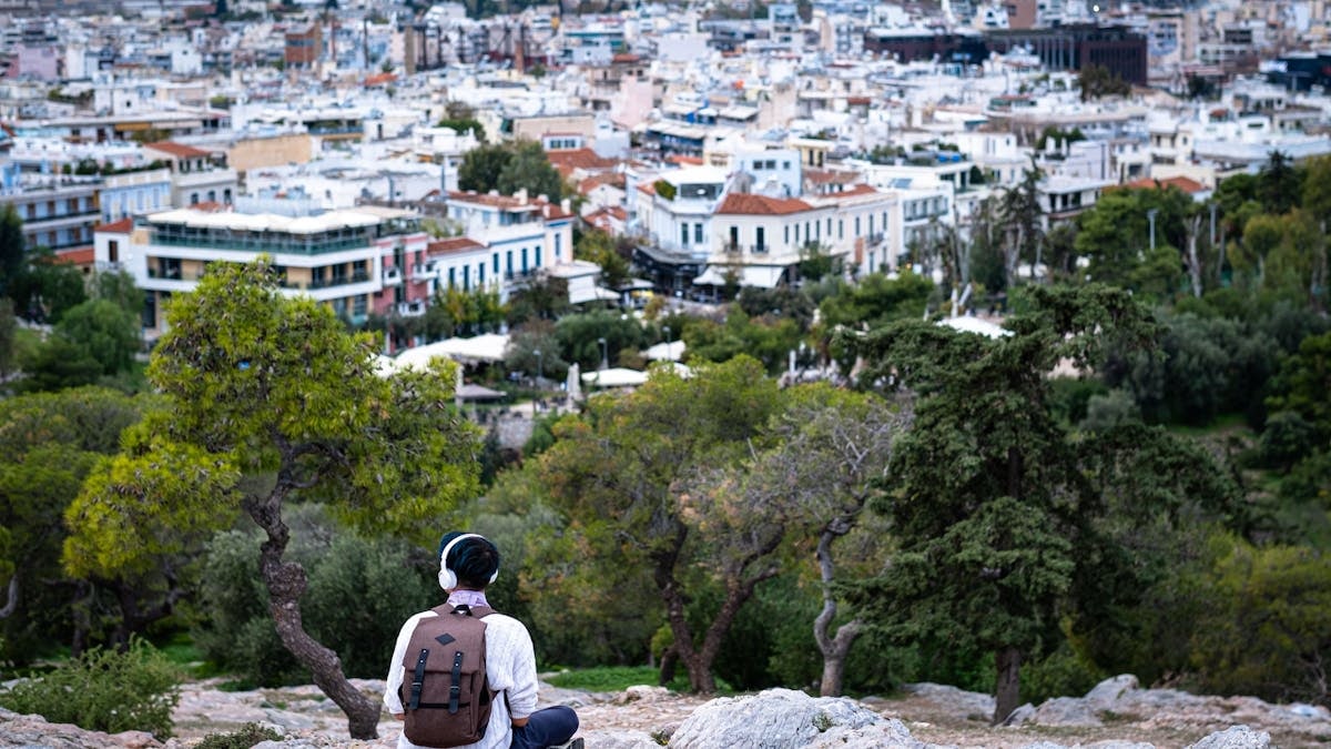 A person with a backpack and headphones overlooking a vibrant urban landscape from a hilltop.